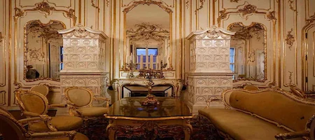Ornate palace sitting room with gilded walls, upholstered furniture and white tiled stoves