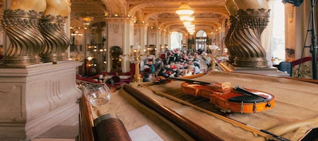 Violin in the foreground of an ornate hall with chandeliers and seated guests
