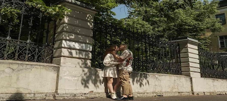Couple kissing beside an ornate iron fence beneath leafy trees